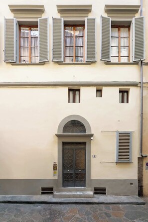 Ein Gebäude mit einem zentralen Eingang, mehreren Fenstern mit Fensterläden und einem gepflasterten Vorplatz.
