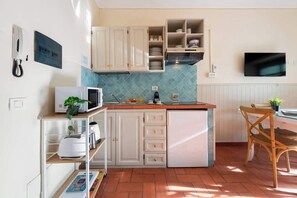Eine kleine Küche mit weißen Schränken, einer Mikrowelle, einem Toaster und einem kleinen Kühlschrank. Es gibt einen Essbereich mit einem Holzbstuhl und einem Tisch.