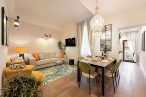 Ein modernes Wohnzimmer mit einer Couch, einem Esstisch, Stühlen, einem Flachbildfernseher und einem großen Fenster mit Vorhängen.