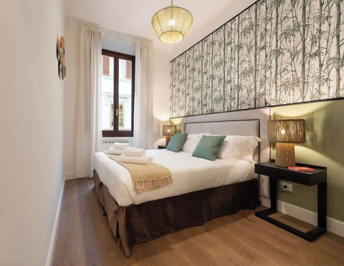 A bedroom with a bed, bedside lamps, a nightstand, a window with curtains, and a wall with a bamboo pattern.
