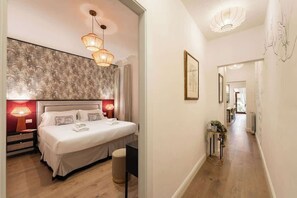 Ein Hotelzimmer mit einem großen Bett, zwei Nachttischlampen, einem Nachttisch, einem kleinen Hocker und einem Flur, der zu einem weiteren Zimmer führt.