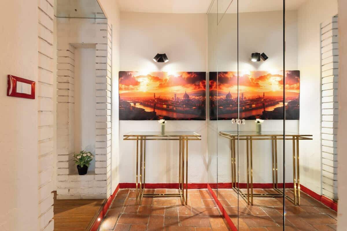 Un couloir avec une cloison en verre, un miroir à cadre rouge, une table en verre et un tableau représentant un paysage urbain.