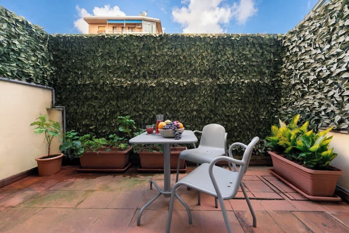 Un patio con tavolo e sedie, circondato da piante in vaso e un muro verdeggiante.