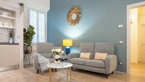 Ein Wohnzimmer mit grauem Sofa, einem kleinen runden Tisch mit Getränken, einer Lampe und einem Wanddekor.