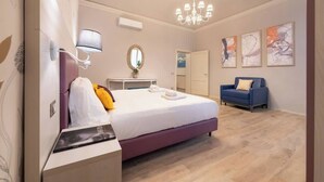Ein Hotelzimmer mit Bett, Schreibtisch, Sessel, Sofa, einer Kronleuchter und Kunstwerken an den Wänden.