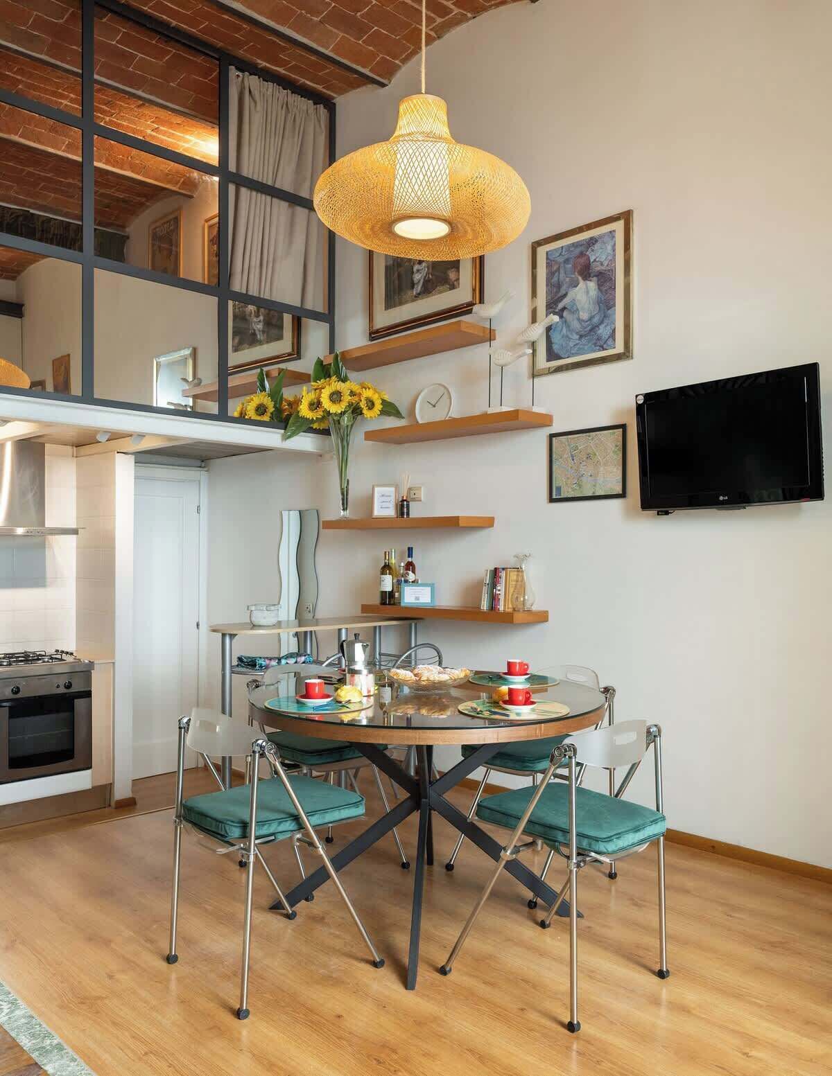 A modern kitchen with a round dining table, green chairs, a hanging light fixture, and a TV mounted on the wall.