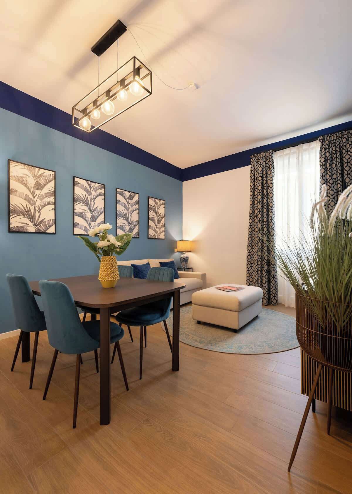 A modern living room with a dining area, featuring a blue accent wall, a wooden dining table, blue chairs, a sofa, a lamp, and a large plant.