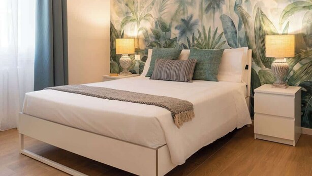 Un dormitorio con una cama, dos mesitas de noche, un cabecero y un mural en la pared, además de una ventana con cortinas.