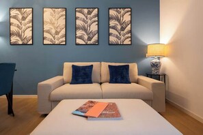 Een kamer met een beige bank, twee blauwe kussens, een klein tafeltje met een lamp, en een muur met vier bladmotieven in lijstjes.