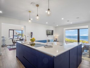 A modern kitchen with a central island, pendant lighting, a large window with a view of the sea, and a flat-screen TV mounted on the wall. - Brentwood/Luxury Ocean View Retreat in Connemara (Renvyle, Connemara)