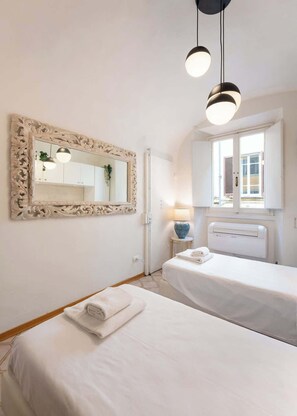 Ein Hotelzimmer mit zwei Betten, einem großen, verzierten Spiegel, einem Fenster mit Jalousien, einer Nachttischlampe und einem Heizkörper.