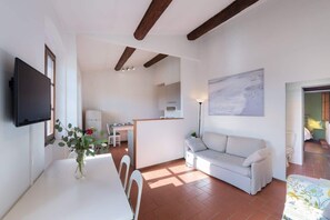 A modern living room with a white sofa, a dining table with chairs, a flat-screen TV mounted on the wall, and a kitchen area in the background. - Pilastri Lorenz Rooftop with 2 Terraces Duomo View (Firenze)
