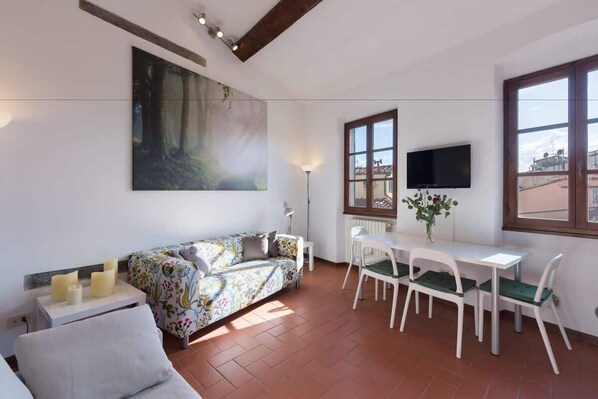 Ein modernes Wohnzimmer mit einer Couch, einem Esstisch mit Stühlen, einem an der Wand befestigten Fernseher und einem großen Fenster.