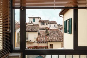 Blick durch ein Fenster mit Jalousien, der eine Straße mit Häusern mit Terrakotta-Dachziegeln und einen Balkon mit Geländer zeigt.