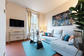 Ein Wohnzimmer mit einer Couch, einem Couchtisch, einem an der Wand montierten Fernseher und einem großen Fenster mit Vorhängen.