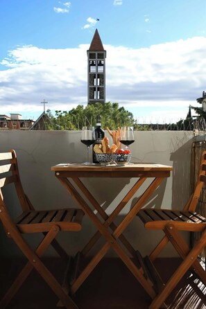 Una mesa de madera con sillas, una botella, vasos y un cuenco con pan y tomates en una azotea con vista a una torre de iglesia.