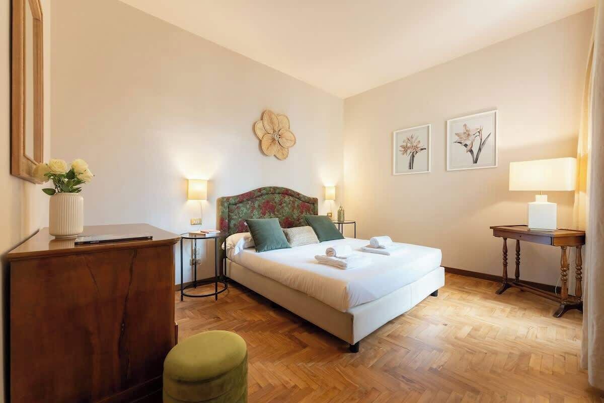 A hotel room with a bed, bedside tables, a desk, a chair, and artwork on the walls.