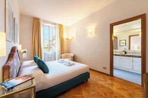 A hotel room with a bed, a chair, a table with a magazine, a window with curtains, and a view of the kitchen. - Antonella Elegant 3-Bedroom in Prime Location (Firenze)