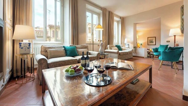 Un salón espacioso con sofá, sillones, una mesa de centro con frutas y copas de vino, y vistas de la ciudad a través de amplios ventanales.