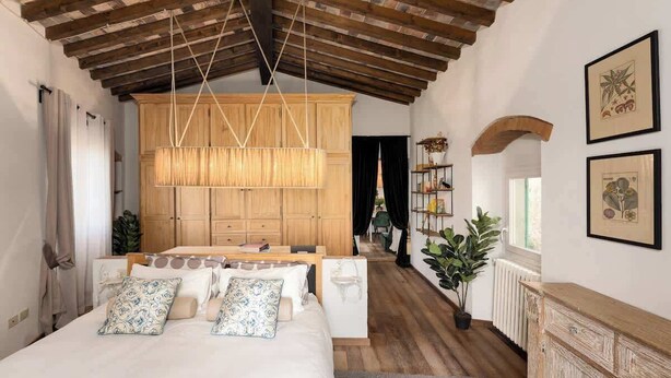 Quarto com cama, vigas de madeira no teto, luminária pendente, guarda-roupa de madeira e uma janela.