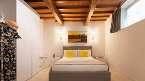 A bedroom with a bed, a nightstand, a wall-mounted lamp, a painting, and a window with curtains. - Casa Falorni, 1 - Raffaello - Bilocale (Greve in Chianti)