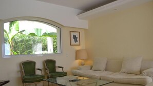Un salon avec une table basse en verre, deux fauteuils verts, un canapé beige, une lampe et une fenêtre donnant sur un paysage verdoyant.