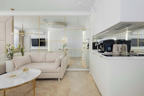 Eine moderne Küche mit Backofen, ein Essbereich mit weißem Tisch und einer Couch, und ein Wohnzimmer mit beiger Couch und Couchtisch.