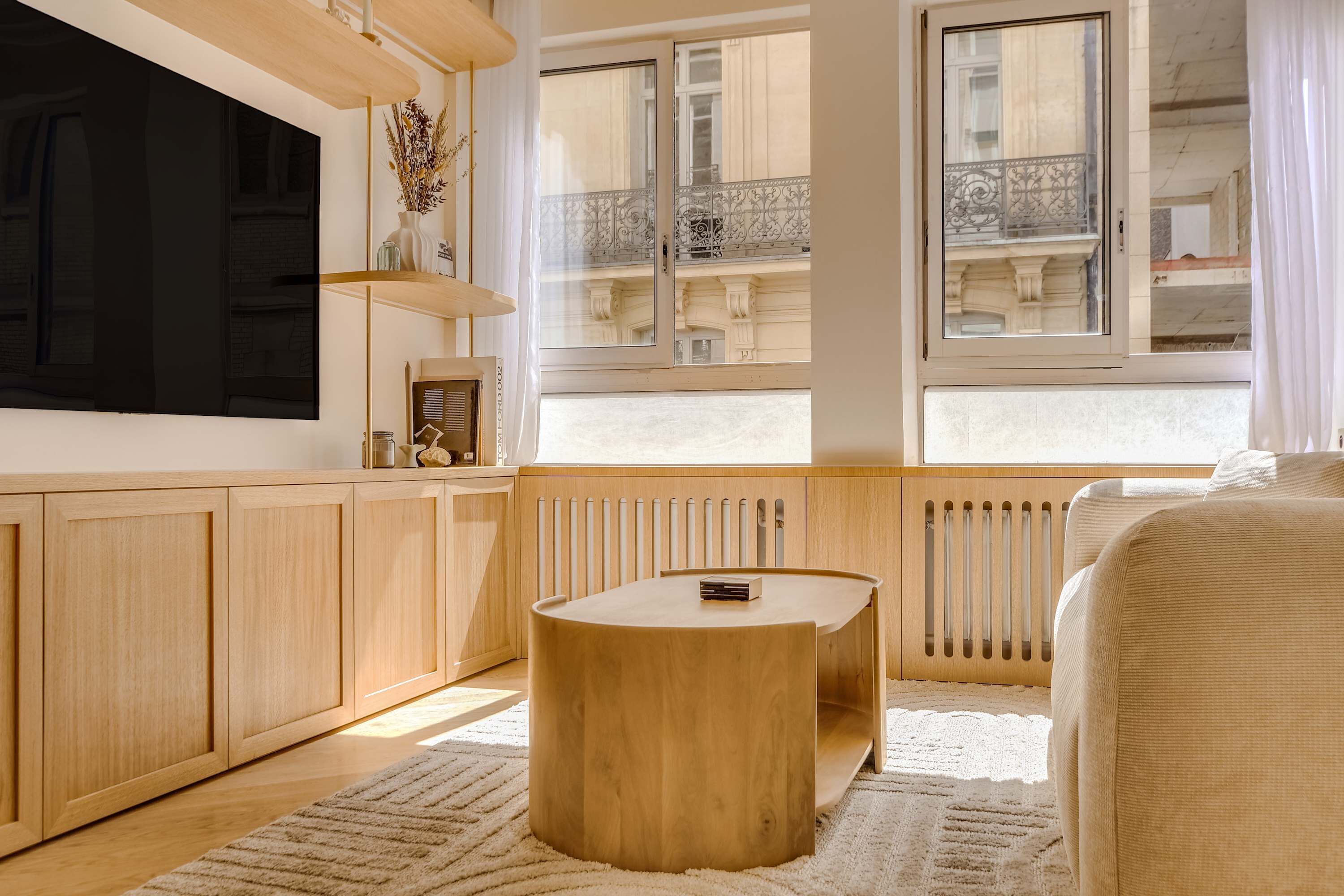 Une chambre moderne avec un grand lit, une table ronde en bois, un téléviseur et une vue sur un immeuble par la fenêtre.