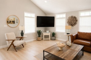 A living room with a flat-screen TV, a brown leather couch, a round mirror, and a coffee table with decorative items.