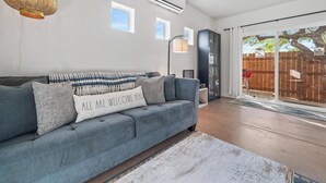 Ett vardagsrum med en grå soffa, en vit kudde med text på, en hög golvlampa och utsikt över ett trästaket genom en glasdörr.