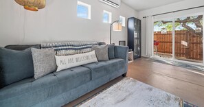 Ein Wohnzimmer mit grauem Sofa, einem weißen Kissen mit Text, einer hohen Stehlampe und Blick auf einen Holenzaun durch eine Glastür.
