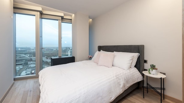 Ein Schlafzimmer mit einem großen Bett, einem Nachttisch, einem kleinen Tisch mit Pflanze und Blick auf die Stadt durch große Fenster.