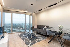 A modern living room with a glass coffee table, grey sofa, and a dining table with chairs. - Stylish 2 Bed Flat in the Heart of Olympic Park (London)