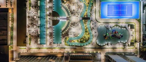 A rooftop pool area with a winding water feature, seating areas, and a tennis court.