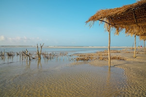 Uma praia com águas cristalinas e rasas, uma construção de palha e uma linha de costa distante.