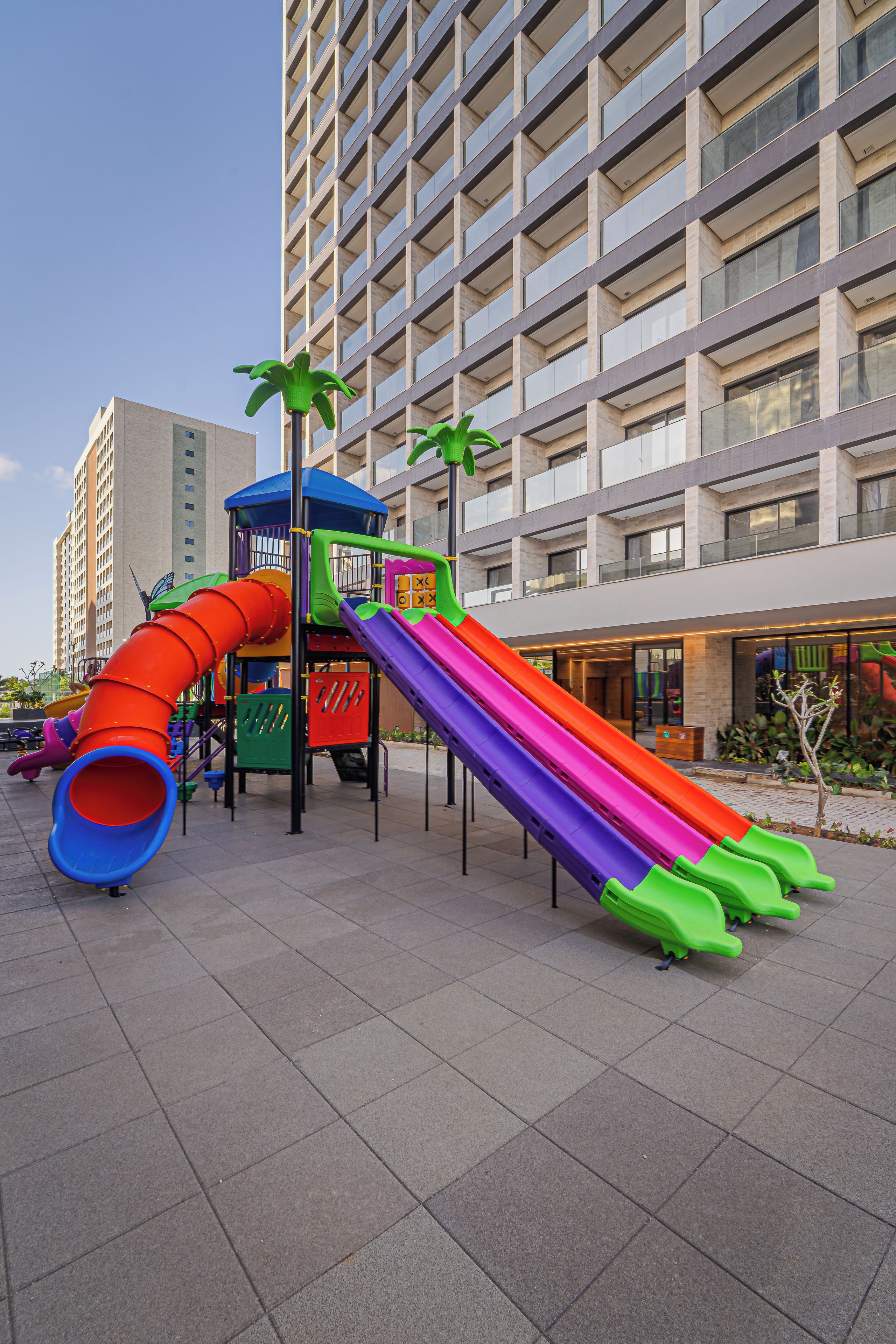 A playground with multiple slides and a climbing structure, located in front of a modern building with balconies.