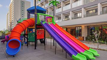 A playground with multiple slides and a climbing structure, located in front of a modern building with balconies.