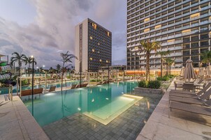 Área de piscina de hotel com céu claro, palmeiras e edifícios modernos ao fundo.