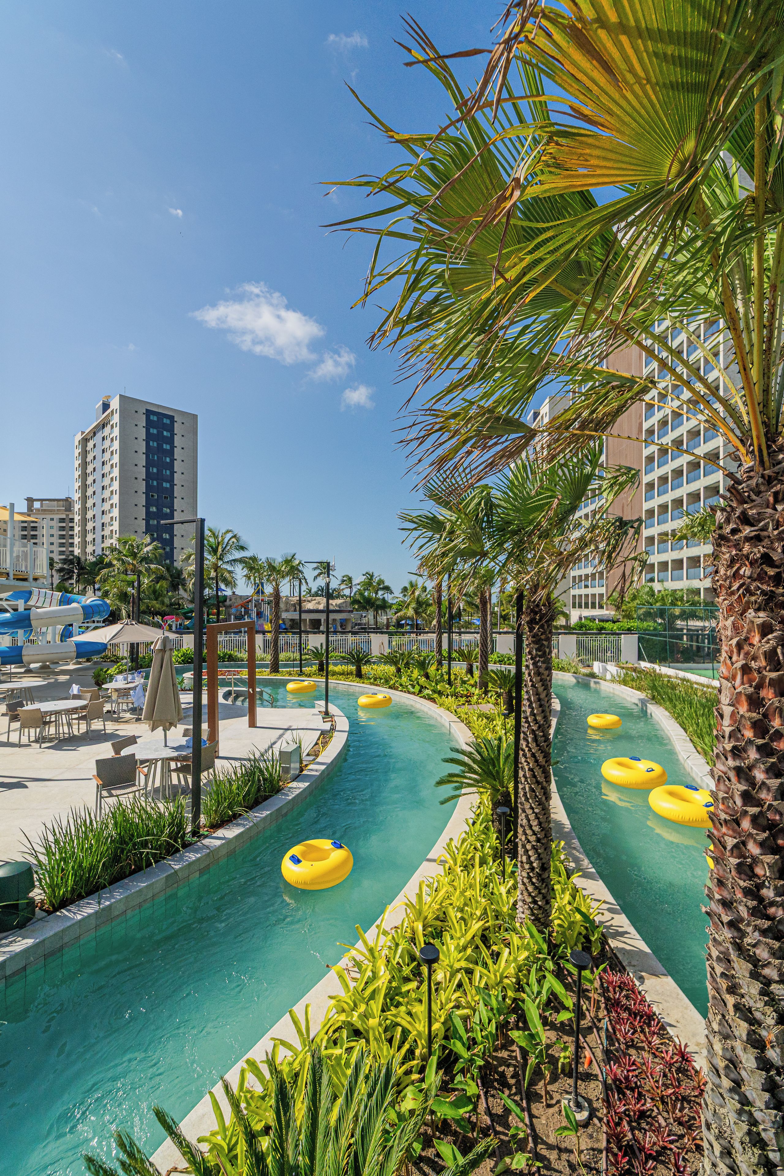 Zona de piscina con varias pistas, palmeras y un edificio al fondo.