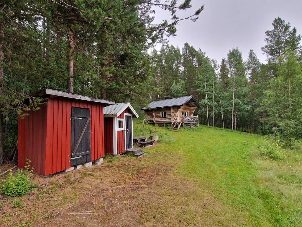 A red cabin with a small porch and a wooden cabin in a forested area.