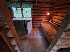 A log cabin interior with a kitchen area, a bench, and a window with curtains.