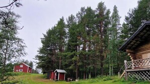 A rustic cabin with a wooden deck, surrounded by tall pine trees. - Serene 1-bedroom lakeside cabin at the arctic circle  perfect for a cozy getaway (Jokkmokk)