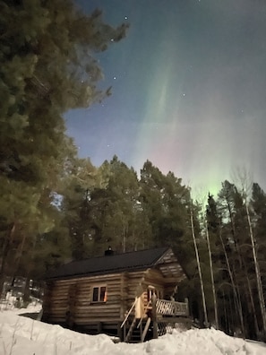 A log cabin with a porch, surrounded by snow and trees, under a night sky with visible stars.