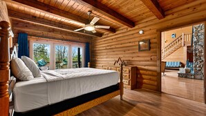 Un dormitorio de cabaña de madera con cama, ventilador de techo, cómoda y una escalera que lleva a una chimenea de piedra.