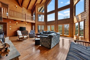 A spacious living room with a large sectional sofa, a wooden ceiling, and large windows offering a view of the outdoors.