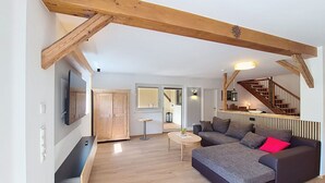 Una sala de estar con sofá, televisión, vigas de madera, una escalera y un pasillo.
