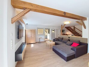 Ein Wohnzimmer mit Sofa, Fernseher, Holzbalken, einer Treppe und einem Flur.