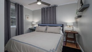 A bedroom with a bed, a ceiling fan, two bedside tables with lamps, a nightstand, and a window with curtains. - Oceanfront Boutique Retreat~Hot Tub~Woodstove (Hearts Delight-Islington)