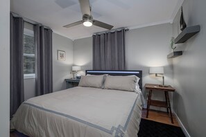 A bedroom with a bed, a ceiling fan, two bedside tables with lamps, a nightstand, and a window with curtains.