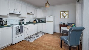 A compact kitchen with white cabinets, a microwave, a stove, and a refrigerator. There is a dining table with chairs and a patterned rug. - Oceanfront Boutique Retreat~Hot Tub~Woodstove (Hearts Delight-Islington)